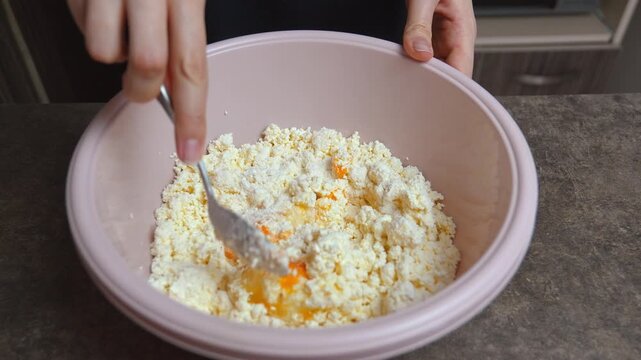 Female hands mixing cottage cheese, egg yolk, and flour with a spoon in a bowl. Homemade recipe for preparing sweet curd dough for syrniki, pancakes, or a delicious cheesecake