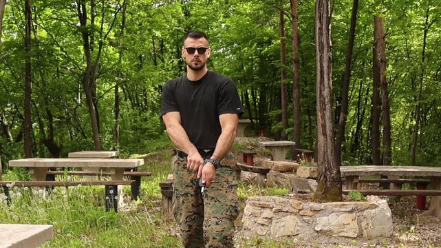 Serious man wearing tactical camouflage pants and sunglasses cocking and aiming an airsoft handgun in a ready to fire position during outdoor shooting training in a dense forest setting.