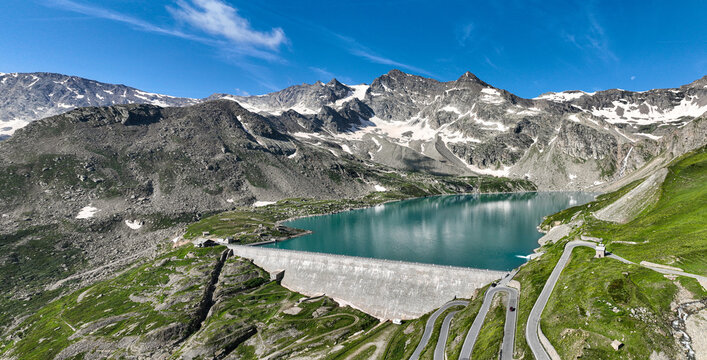 Aerial view of Serru Lake and its massive dam surrounded by rugged alpine peaks with snow patches and winding mountain roads in Ceresole Reale, Piedmont, Italy.