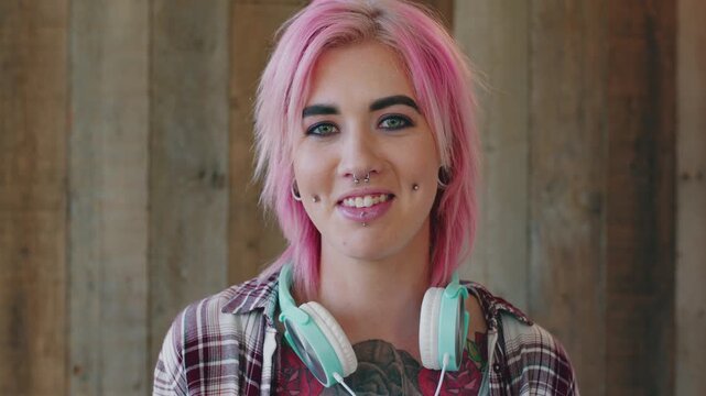 Face, woman and punk style by wood background with tattoo, headphones and pink hair for edgy aesthetic. Happy, person and alternative fashion with audio tech, piercings and makeup for individuality.