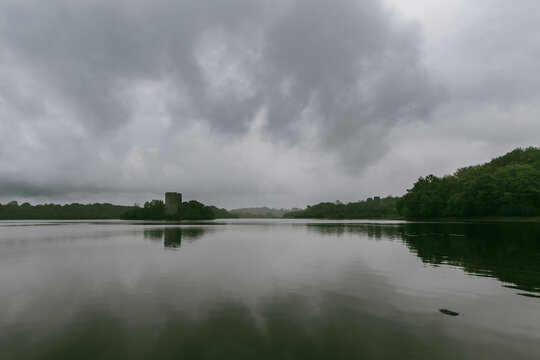 Cloughoughter Castle rises from Lough Oughter in County Cavan, Ireland, beneath a dramatic clouded sky. A tranquil medieval tower house reflected in the calm lake.