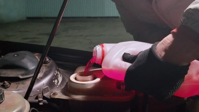 Male mechanic with gloves pouring antifreeze into the cooling system expansion tank of a car with an open hood during vehicle maintenance in a service garage