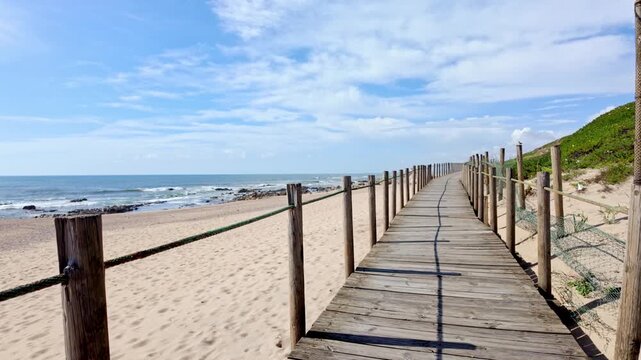 A rustic wooden walkway stretches across golden dunes toward the horizon under a bright sky. Warm sunlight highlights the weathered wood texture and creates long shadows on the path.