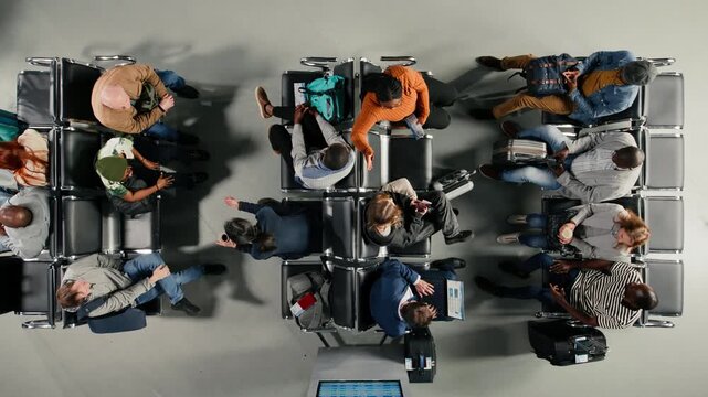 Top down view of tourists in airport lounge boarding for departing airplane flights, standing up from chairs. Passengers leaving airport terminal, going to boarding gate with trolleys, high angle shot