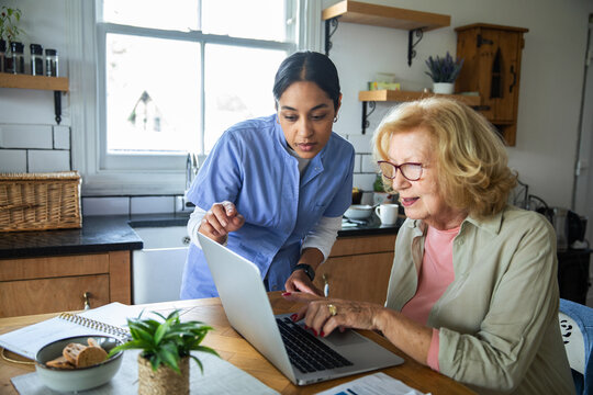 Caregiver helping senior woman with laptop at home kitchen
