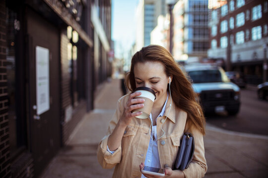Smiling woman drinking coffee and using smartphone on city street
