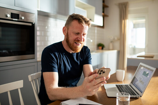 Smiling bearded man using smartphone and laptop in home kitchen