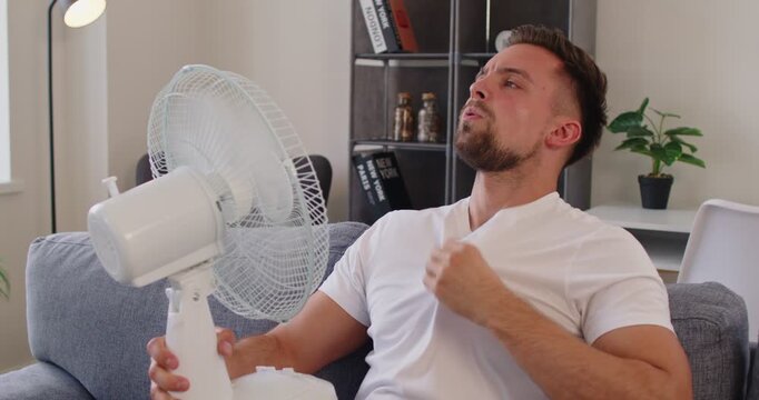 At home, a man on a couch faces heat, using a fan for cooling as hot air lingers through summer, tugging his shirt collar and wiping sweat while seeking relief. Man cools down during heatwave.