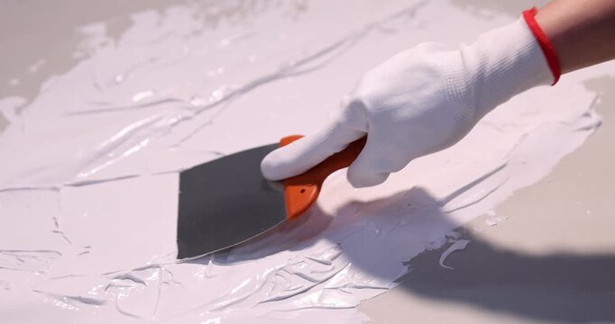Woman in protective gloves applying white plaster with putty knife onto grey surface, closeup