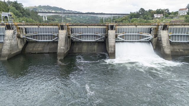 Aerial view of the Velle Dam with water flowing through a spillway gate on the Mino River surrounded by green hills and a bridge in Ourense, Ourense, Spain.