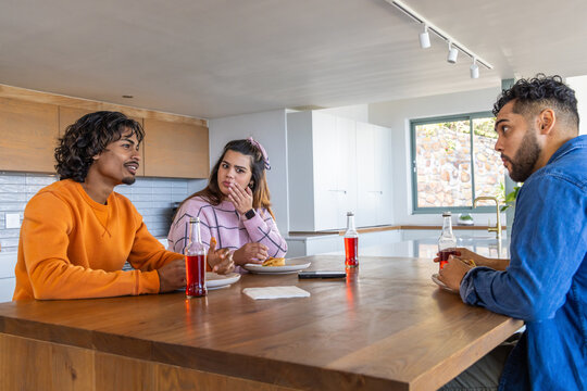 Diverse friends chatting at wooden island in kitchen, wearing colorful clothes, sipping red soda