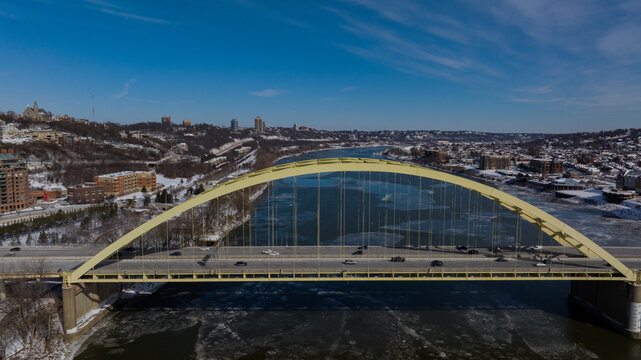 Aerial view of the yellow Daniel Carter Beard Bridge spanning the icy Ohio River with snow-covered hills and urban buildings under a clear blue sky in Cincinnati, Ohio, United States.
