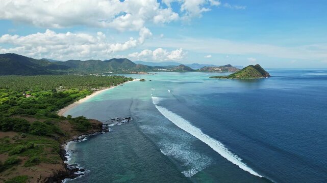 Aerial drone footage of Kertasari surfing spot bay, in West Sumbawa, Indonesia, with crystal clear turquoise and blue water, white sand and long waves with reef, vegetations, small island and village