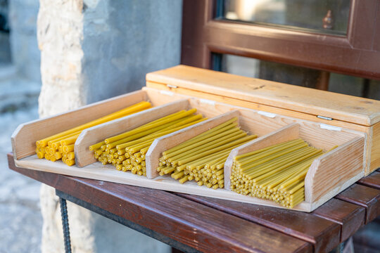 Yellow beeswax church candle neatly arranged in compartment of wooden display box on table near stone wall and door, religious faith concept.
