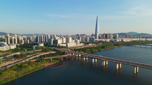 Aerial rising revealing Seoul city skyline featuring iconic Lotte World Tower skyscraper and Jamsil Bridge traffic crossing Han River under clear blue sky in South Korea urban landscape