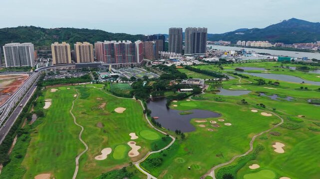 Aerial forward flight over Macau International Golf Course showcasing lush fairways, sand bunkers, water hazards, pond and mountain backdrop with residential skyline