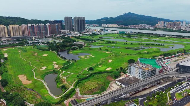 Aerial flyover over Macau International Golf Course revealing lush green fairways, sand bunkers, water hazards and scenic mountain backdrop with city skyline