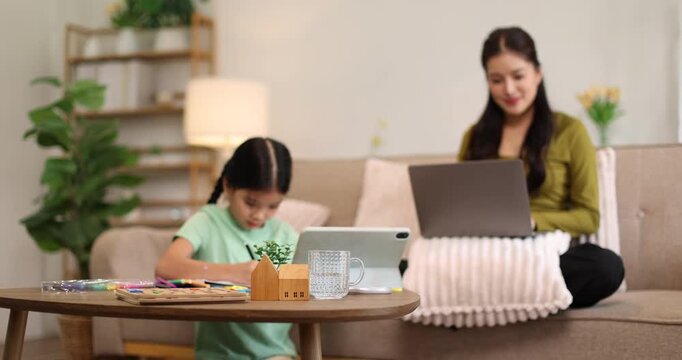 Mother working on laptop at coffee table while daughter focuses on drawing and learning nearby, illustrating remote work, distance education and family bonding in a cozy living room
