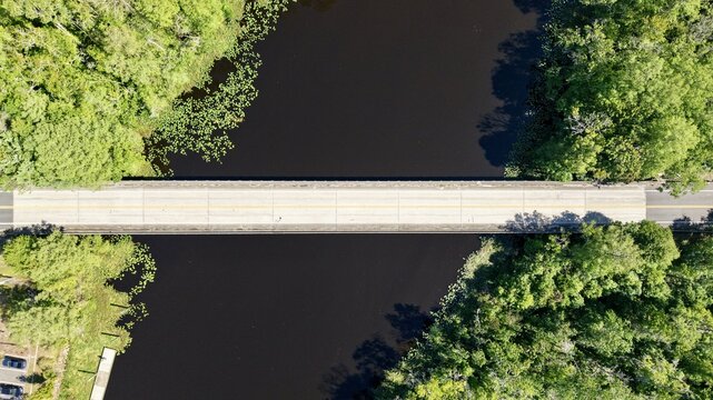 Aerial view of Sixmile Creek bridge crossing dark water surrounded by dense green forest and lily pads in St. Johns County, Florida, United States.