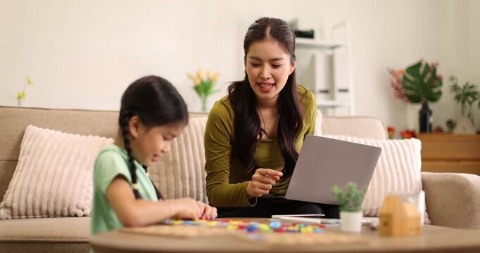 Asian mother smiling while working on a laptop on the sofa as her young daughter plays educational games at the coffee table, capturing remote work and family bonding