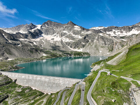 Aerial view of Serru Lake and its massive dam surrounded by winding mountain roads and snow-capped peaks in the Graian Alps, Ceresole Reale, Piedmont, Italy.