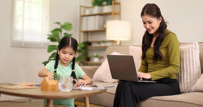 Asian mother sitting on a sofa and focusing on a laptop, working from home while her young daughter is playing with a puzzle game on a low table in the living room, depicting work-life balance
