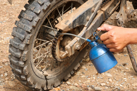 Focused mechanic hand holds blue oil can applying liquid lubricant to metal chain and sprocket of rugged dirt bike wheel during routine offroad motorcycle maintenance outdoors on dirt ground