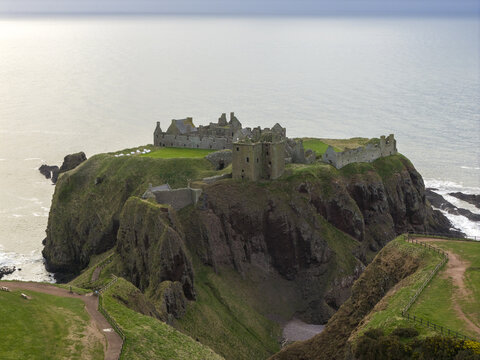 Aerial view of Dunnottar Castle ruins perched on a dramatic cliff overlooking the North Sea with green grass and steep rocky slopes Stonehaven, Scotland, United Kingdom.