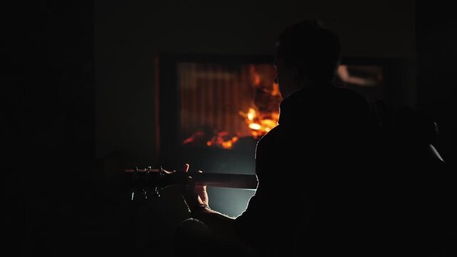 Silhouette of man playing guitar in dark room in front of a burning fireplace, creating a cozy and warm atmosphere, cinematic motion footage.