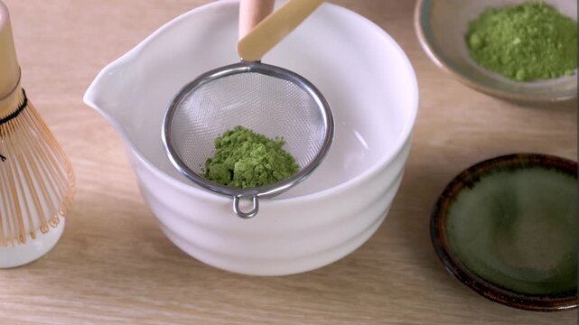 Sifting matcha powder into bowl and whisk on grey table, flat lay
