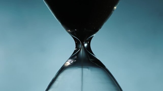 Close up cinematic shot of black grains of sand flowing down in hourglass as time passes against a bright background.