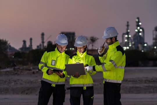 Group of professional petroleum engineers in high-visibility jackets and hardhats working together, using laptop and tablet computers at oil refinery chemical plant industry factory site at twilight.