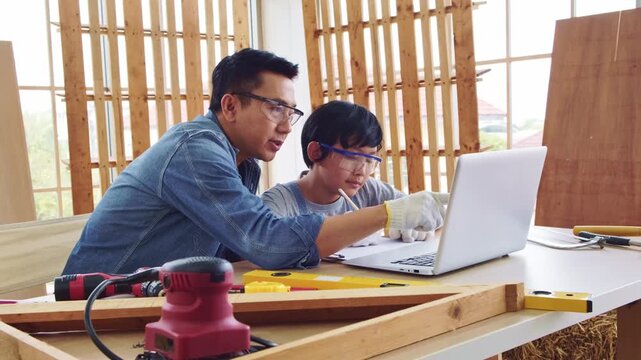 Cheerful Asian carpentry father discusses and teaches his son about construction instruments on laptop at the home workshop studio. Construction work and repairs. Carpentry and family concept.