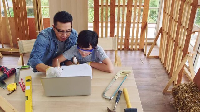 Cheerful Asian carpentry father discusses and teaches his son about construction instruments on laptop at the home workshop studio. Construction work and repairs. Carpentry and family concept.