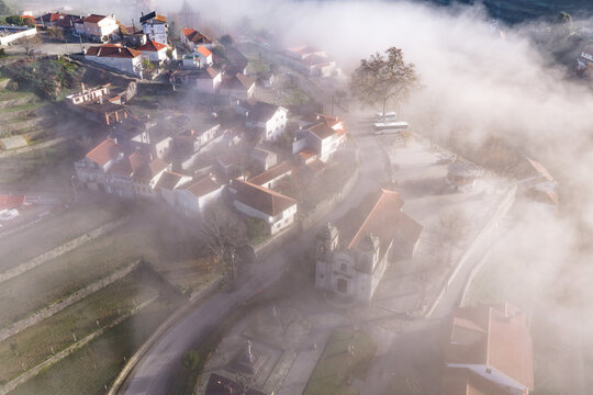 Aerial view of Avo village with red-roofed houses and a historic church partially covered by thick morning fog in Oliveira do Hospital, Coimbra District, Portugal.