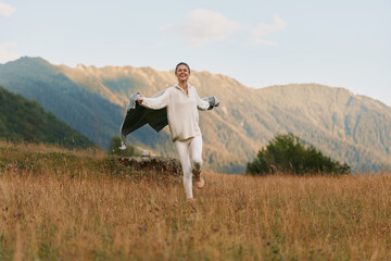 Fototapeta premium Smiling woman in a white blouse dances in a golden field with mountains behind, portraying a carefree outdoor lifestyle, joy, and freedom under bright light and glow.
