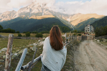 Naklejka premium Young woman with long hair wrapped in blanket stands near wooden fence looks at mountain landscape on sunny day in rural area with dirt road and old house.