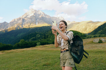 Fototapeta premium Young woman on a hiking trip takes a selfie in a mountain meadow, backpack ready, bright sky, cheerful mood amid vast nature and wildflowers sway nearby under blue sky.