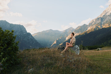 Fototapeta premium Young woman sits on a rock in a meadow with distant mountains, enjoying a peaceful sunny moment.