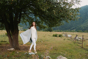 Fototapeta premium A woman in white clothes stands near a tree in a green field with mountains in the background. She holds a light shawl and enjoys the peaceful outdoor lifestyle.