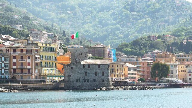 Wide shot of Rapallo Castle and vibrant Ligurian buildings along the waterfront, bathed in daylight. Calm blue water, lush green hills, and an Italian flag create a lively, inviting scene.
