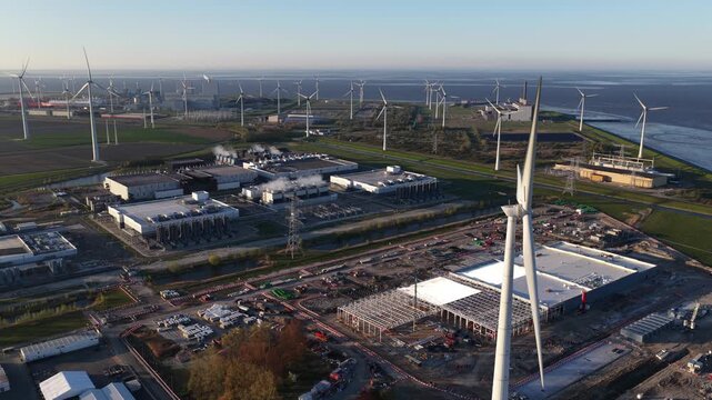 Aerial view of a large data center and technology campus under construction, surrounded by a vast wind farm generating renewable energy for the facility and the power grid near the coastline