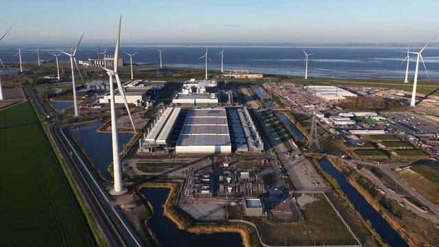 Hyperscale data center complex powered by renewable energy from surrounding wind turbines, showcasing sustainable technology and green energy infrastructure in a coastal industrial area