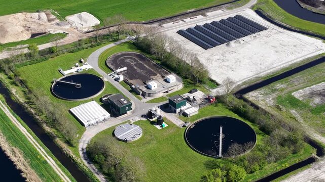 High angle perspective showing a modern sewage treatment facility with circular clarifiers, aeration tanks, and sludge drying beds surrounded by lush green fields and waterways