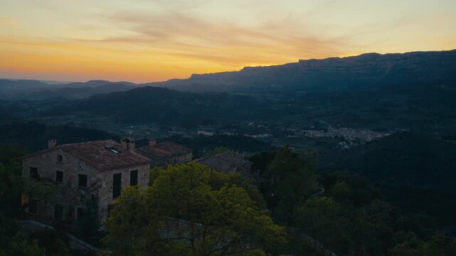 A stone house sits above a darkening valley in Spain as warm sunset color lingers over distant ridges, blending village calm with a wide evening landscape.