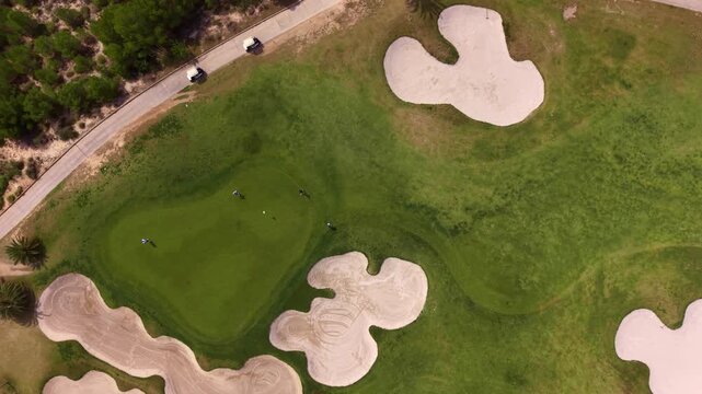 Overhead view of a golf course green surrounded by multiple sand bunkers, with players on the fairway, a cart path, and lush trees bordering the course