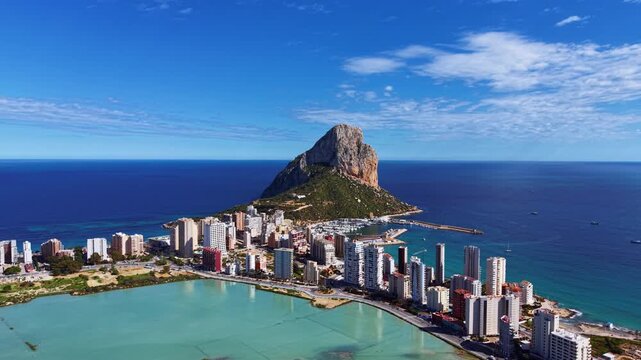 Panoramic view of Calpe, Spain, featuring the iconic Penon de Ifach rock formation rising above the coastal city, marina, turquoise lagoon and blue Mediterranean Sea