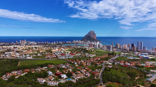 Panoramic view of Calpe, Spain, featuring the iconic Penon de Ifach rock formation rising above the Mediterranean coastline with urban sprawl and blue skies