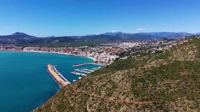 Javea coastal town on Spain's Costa Blanca with turquoise Mediterranean waters, marina filled with sailboats, sandy beach, hillside buildings and mountain backdrop