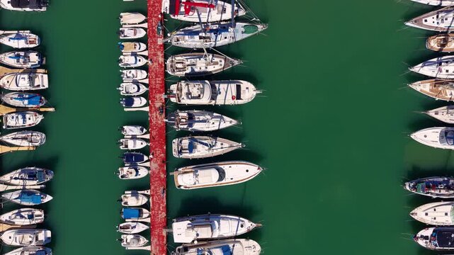 Top-down view of a busy marina with rows of white sailboats and motorboats moored along a red dock pier surrounded by vivid green water